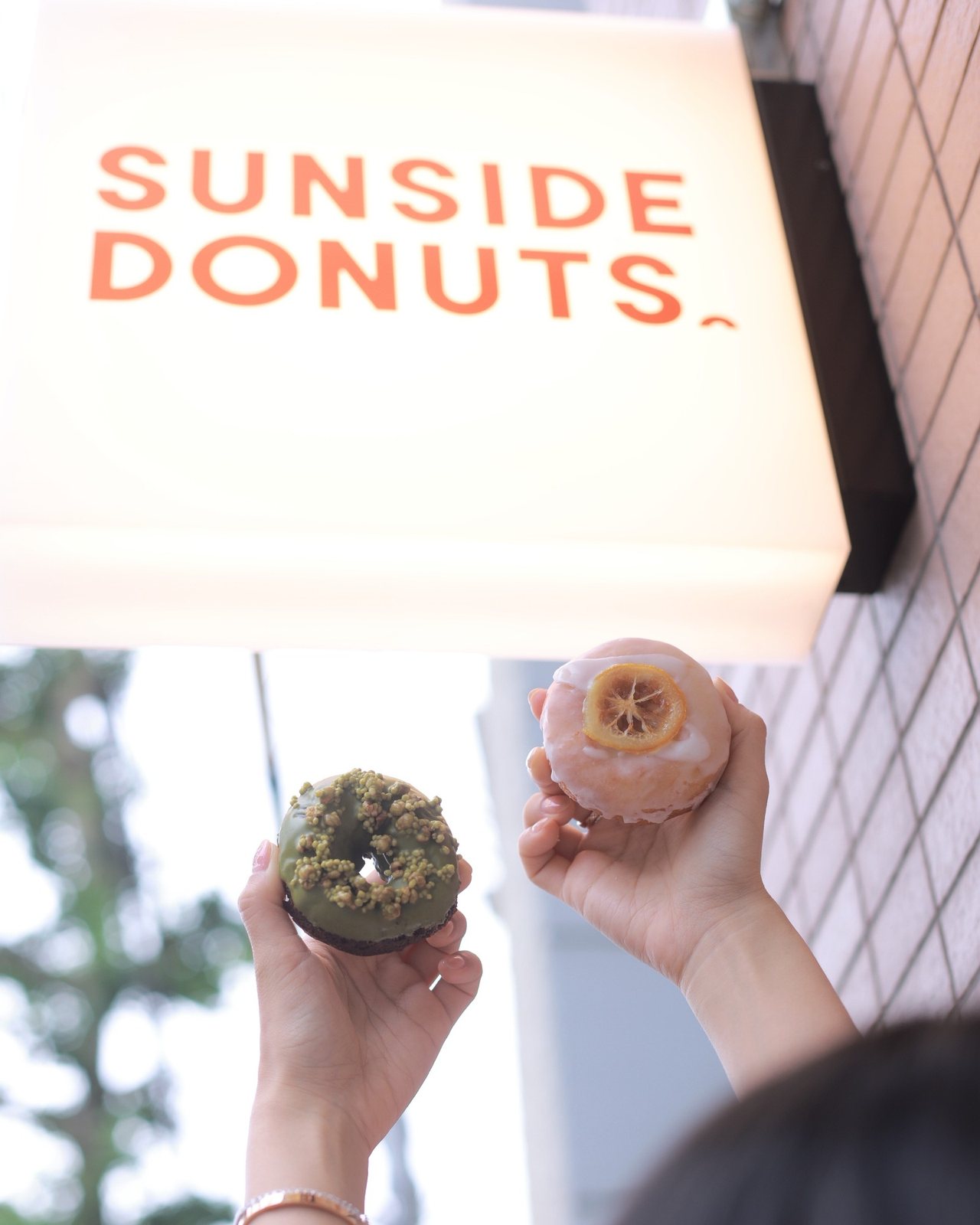 Two donuts held up under a shop sign: matcha and lemon